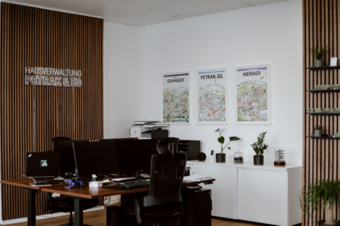 Zwei Schreibtische im Büro der Hausverwaltung Petrak & EG. Sie sind mit zwei Bildschirmen ausgestattet. An der Wand hängen Bilder und das Logo.