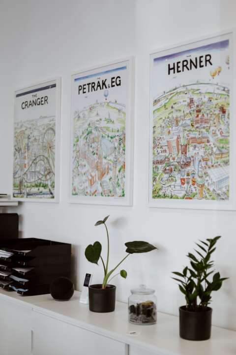 Ein weißer halbhoher Schrank mit Pflanzen, Dokumentenablage und an der Wand drei Bilder von Herne.
